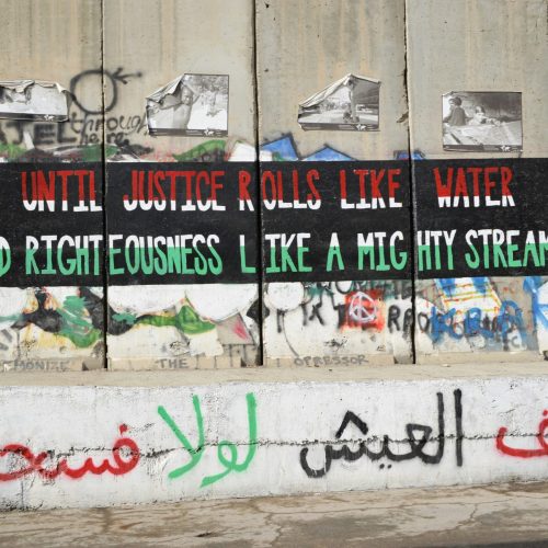 On the huge separation wall in Israel by the West Bank, text reads: "UNTIL JUSTICE ROLLS LIKE WATER AND RIGHTEOUSNESS LIKE A MIGHTY STREAM," alongside other graffiti.