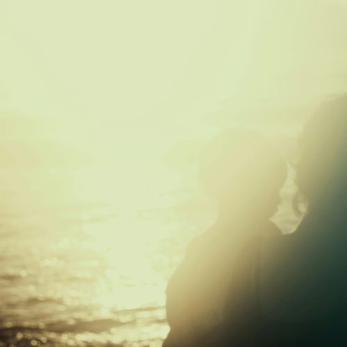 A photo of a translucent, silhouetted woman holding a child, the sea behind them. The photo is bright, overexposed, with a golden-beige tint.