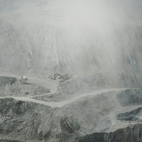 A grey, apocalyptic, hazy scene of mining on a mountainside. Trucks and other equipment sit on one of the mountainside's roads.