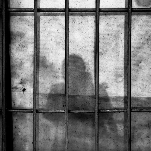 The silhouette of a person who appears to be a woman behind bars, visible through a light-coloured translucent screen. The person may be holding a child or toy. The photo is in greyscale.